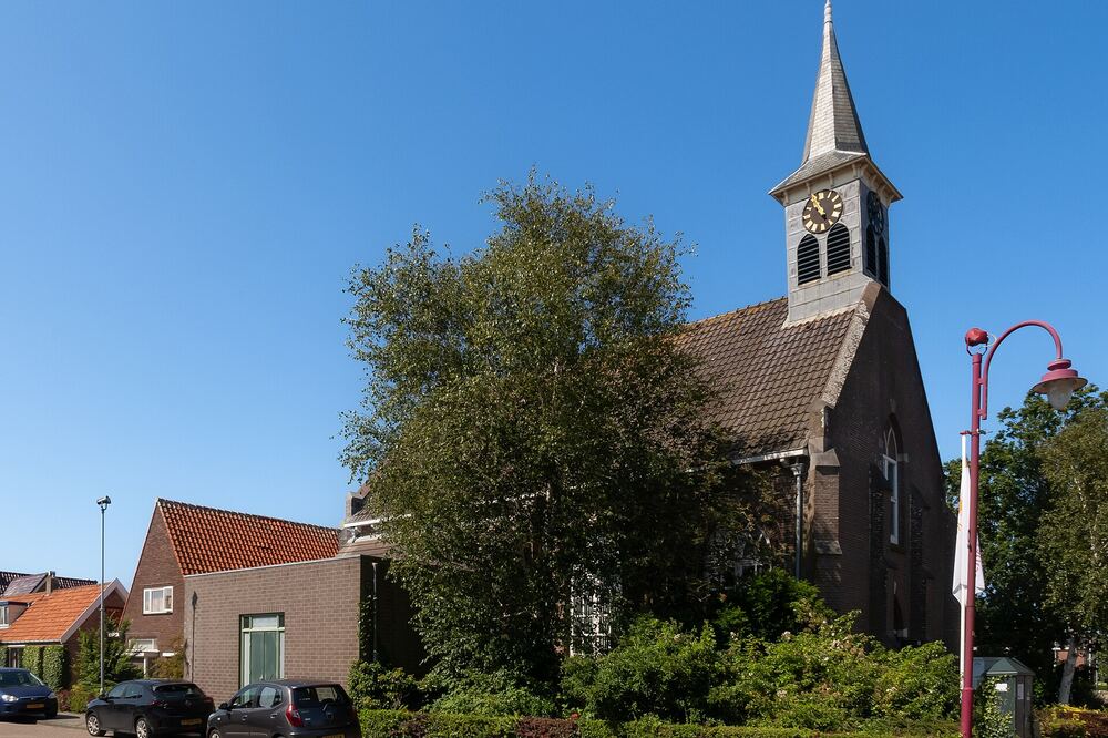 De Ontmoetingskerk in Julianadorp met spitse toren tussen bomen en woonhuizen onder een blauwe lucht