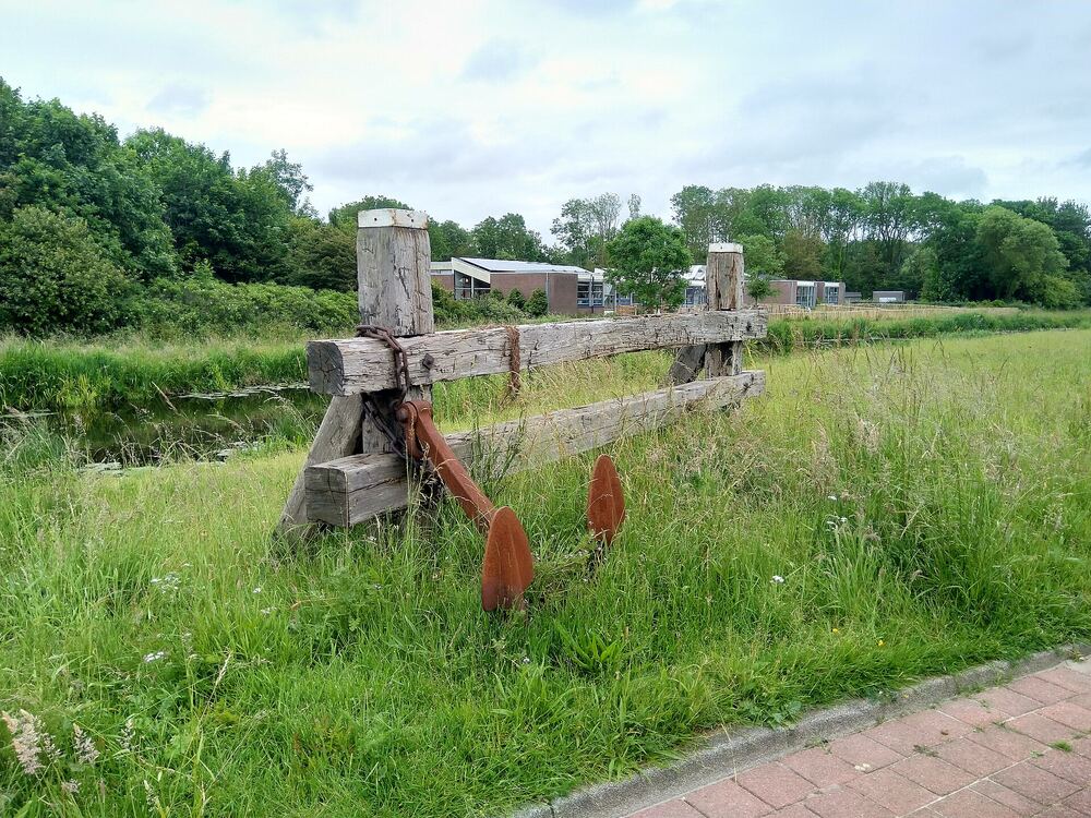 Kunstwerk van meerpalen en een anker in een grasveld langs een wandelpad aan de rand van Julianadorp